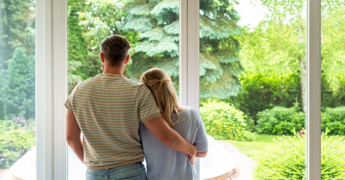A couple standing side-by-side while looking out the tall floor-to-ceiling glass windows leading to their yard.
