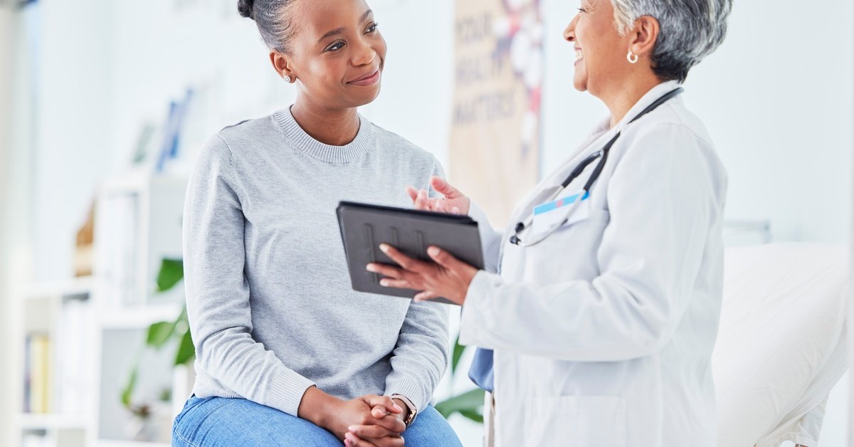 Physician reviews information on tablet with seated patient, both engaged in conversation in clean medical office.