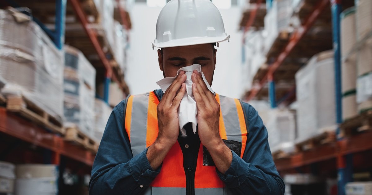 A warehouse worker in a hard hat and orange safety vest sneezes into a tissue while standing between tall storage shelves.