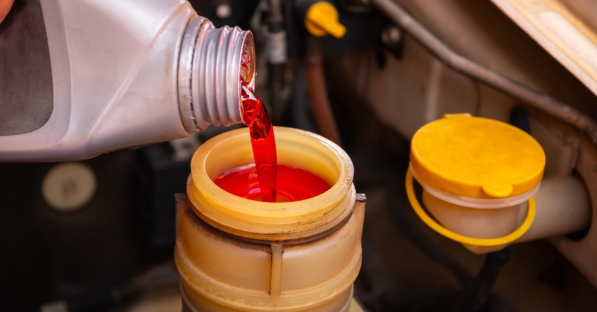 A close-up of a grey plastic container pouring red brake fluid into the yellow fluid reservoir of a car.