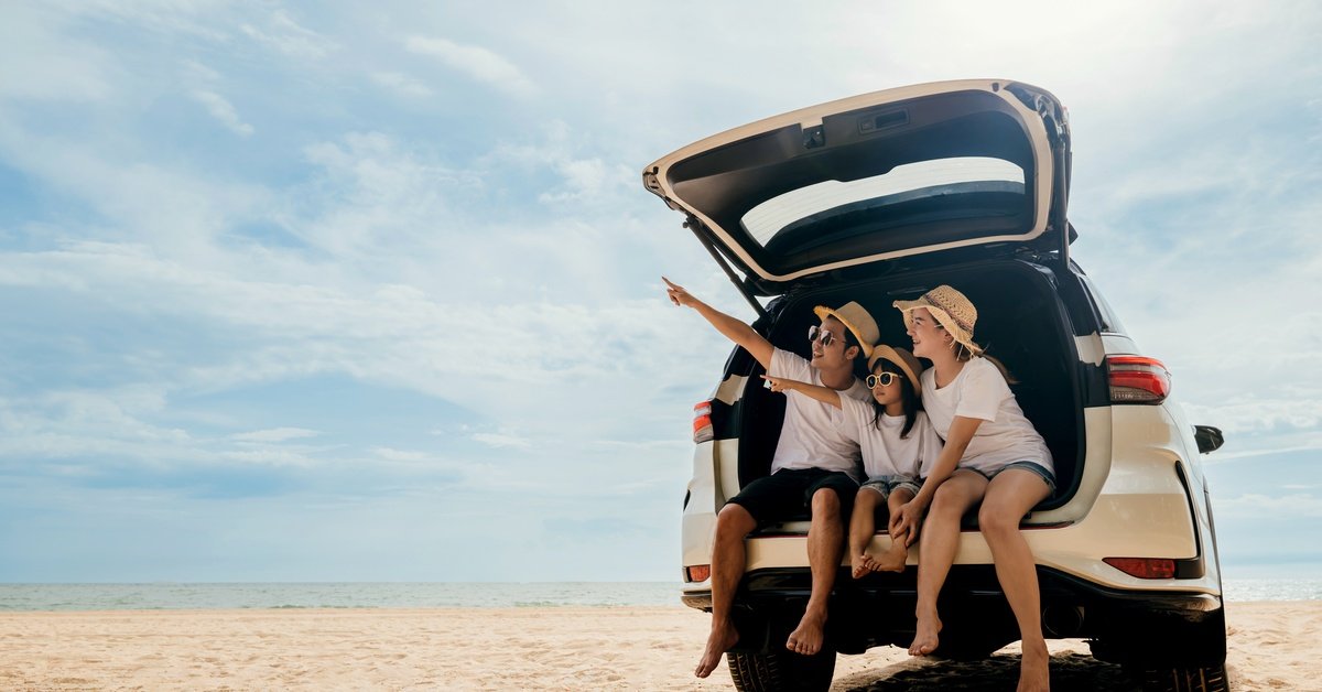 A family sitting in an open SUV trunk on a sandy beach, wearing hats and sunglasses, under a bright blue sky.