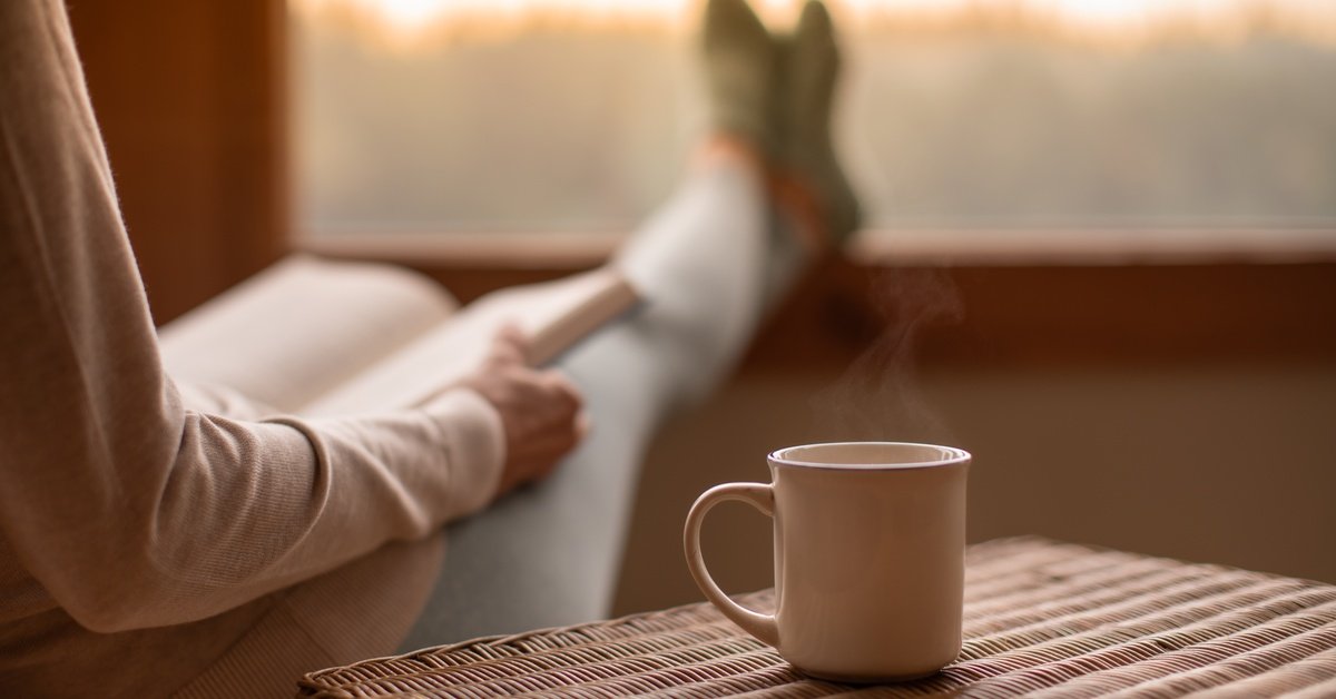 A person relaxing in their home while reading a book. They have a hot drink in a mug on the table next to them.