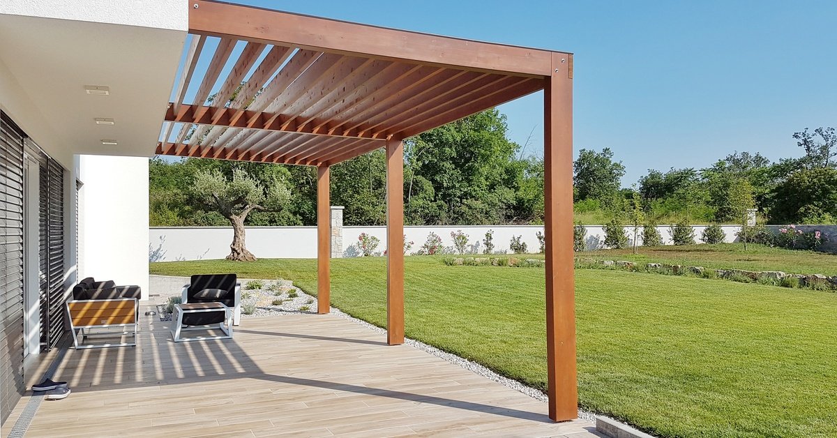 A modern home's patio featuring an attached brown wooden pergola with open vertical slats, overlooking a large flat yard.