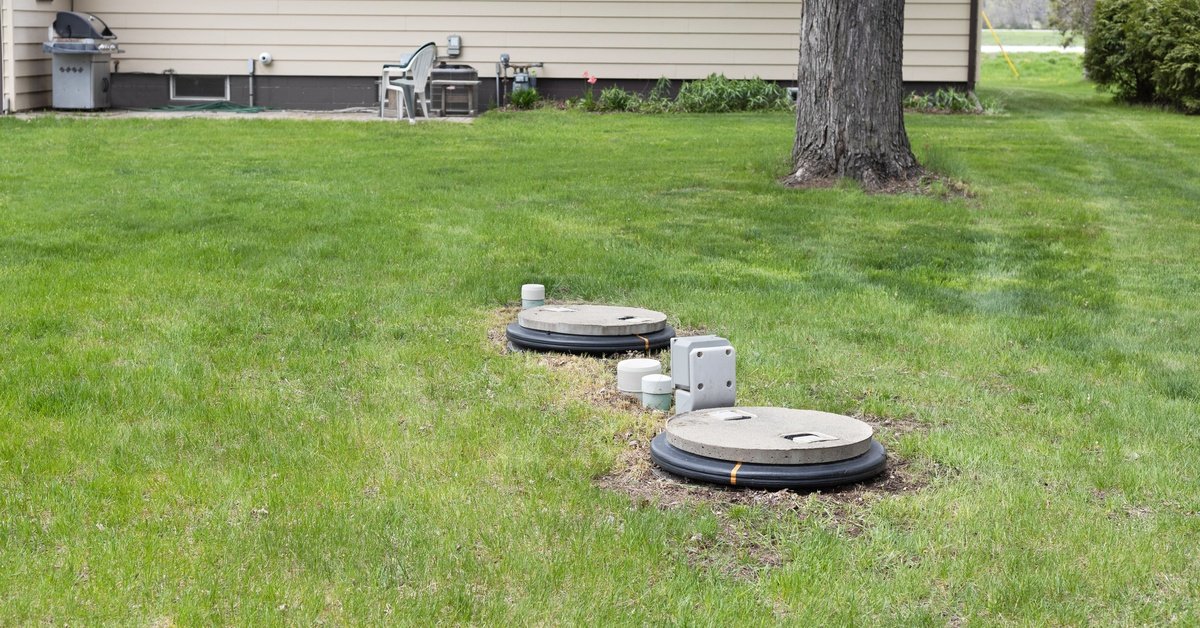 A beige house's backyard that has a septic tank system that is sticking out from the grass of the lawn.
