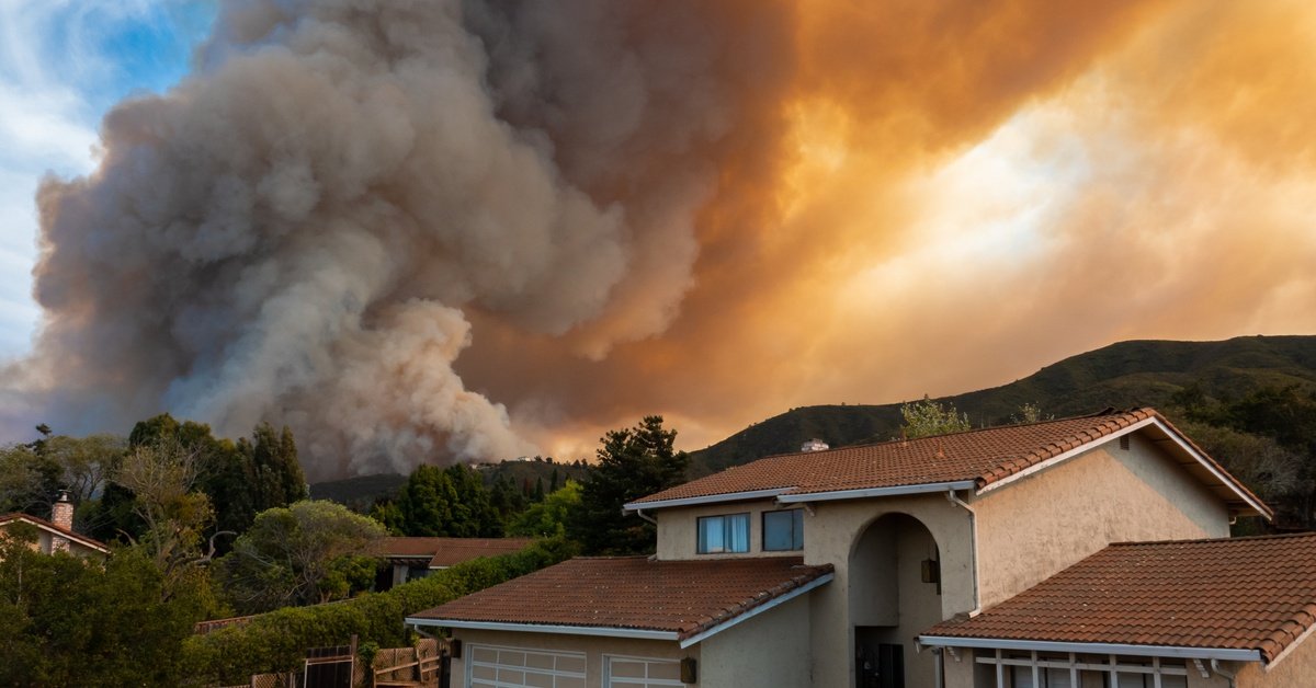 A suburban home with a tiled roof under a massive wildfire smoke plume, an orange sky looming over nearby hills and trees.