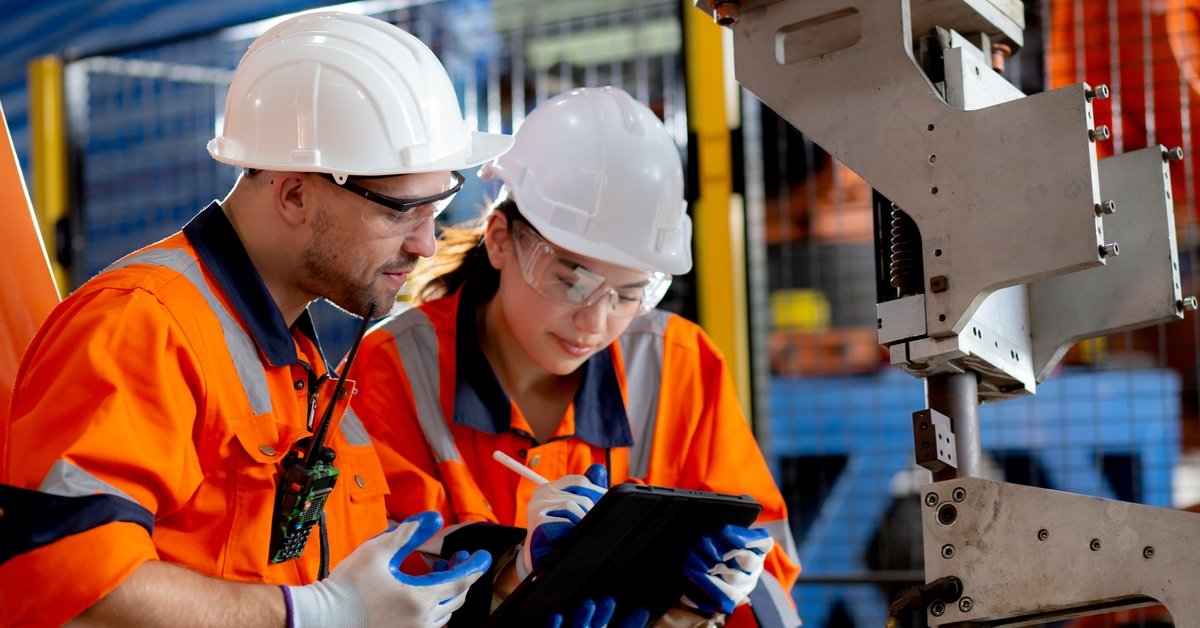 Two industrial workers wearing hard hats. One employee holds a tablet while the other worker writes on it with a pen.
