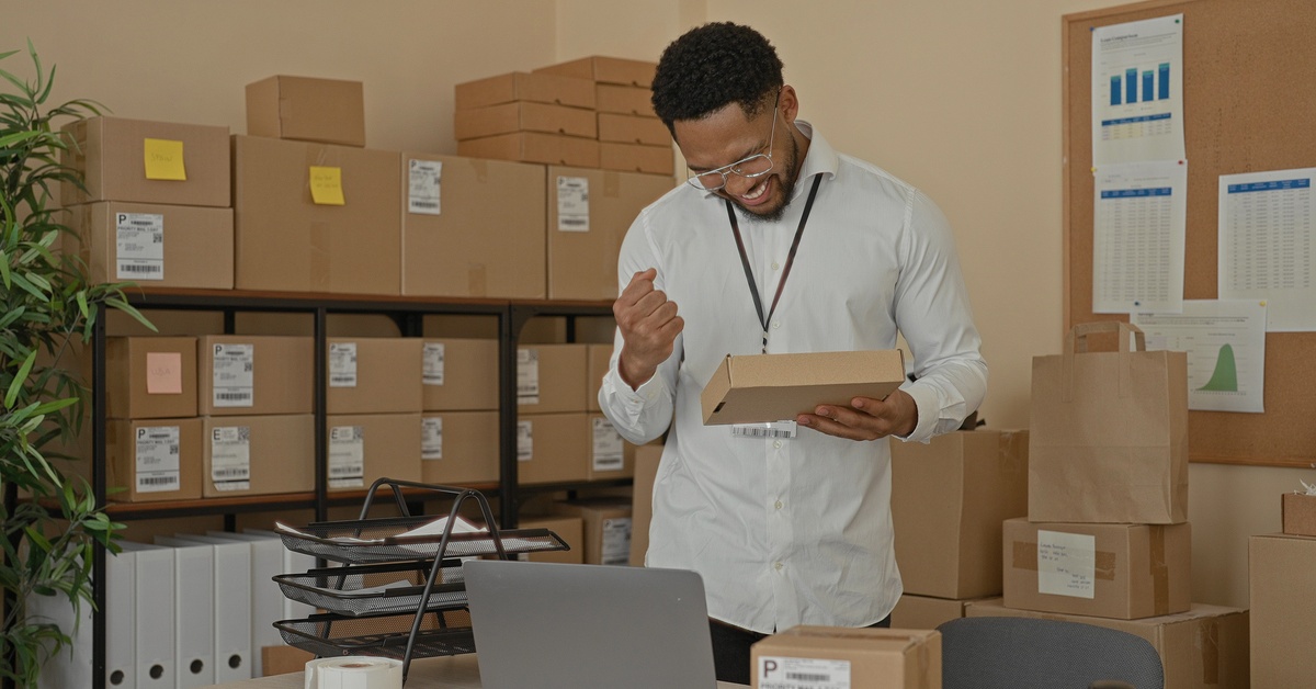 A smiling man is holding a small cardboard box. A shelving unit is behind him with more cardboard boxes.