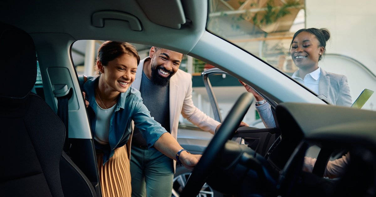 A smiling couple looking into the front seat of a new car while a car salesperson stands just off to the side.