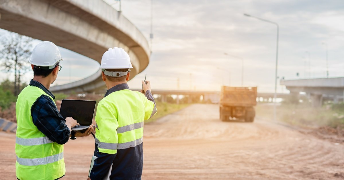 Two construction workers in high-visibility vests assessing a construction site of a new road with a laptop.