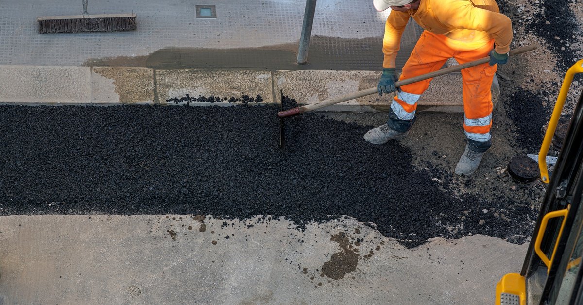 A person in orange safety gear using a rake to spread fresh black asphalt on a paved sidewalk at a roadwork site.