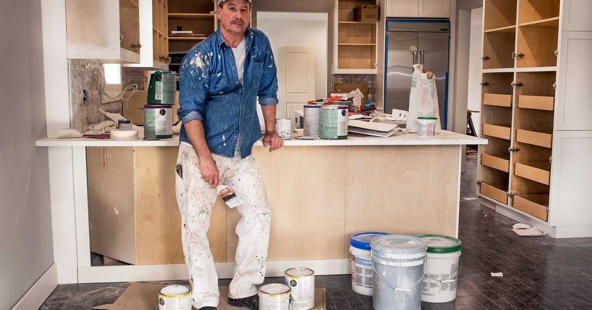 A man wearing paint-splattered clothing is leaning against the countertop in the kitchen. There are paint buckets on the floor.