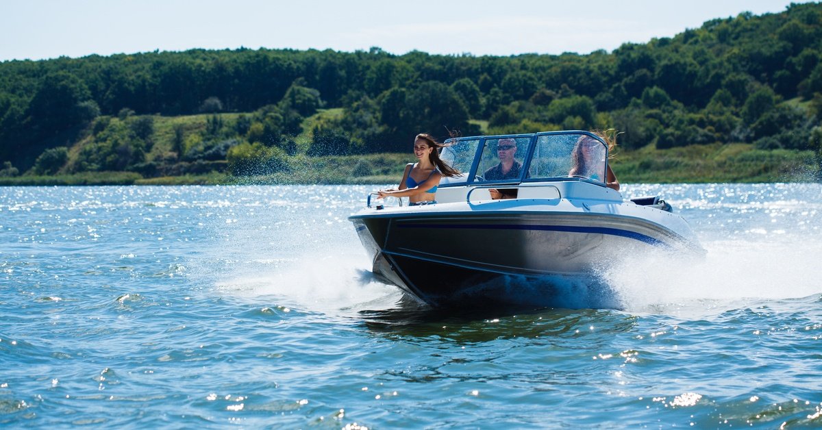 A white and blue motorboat moves across a lake with three people on board. Green wooded hills line the background.