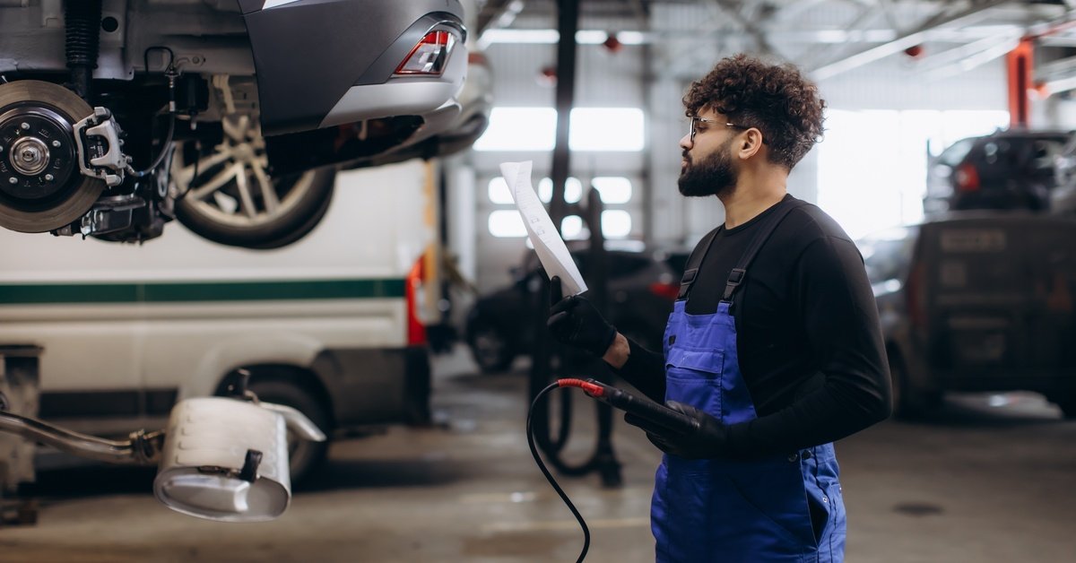 A man wearing a black T-shirt and a blue apron is holding a device that controls the hoist brake. He's looking at a paper.