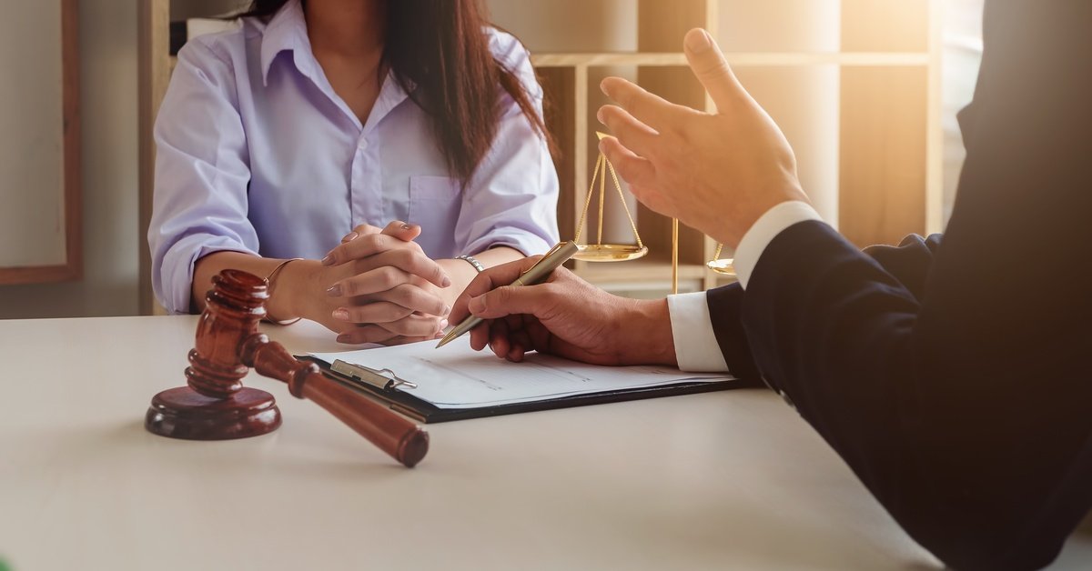 A client sits across from a lawyer discussing legal documents, with a judge's gavel and scales of justice on the desk.