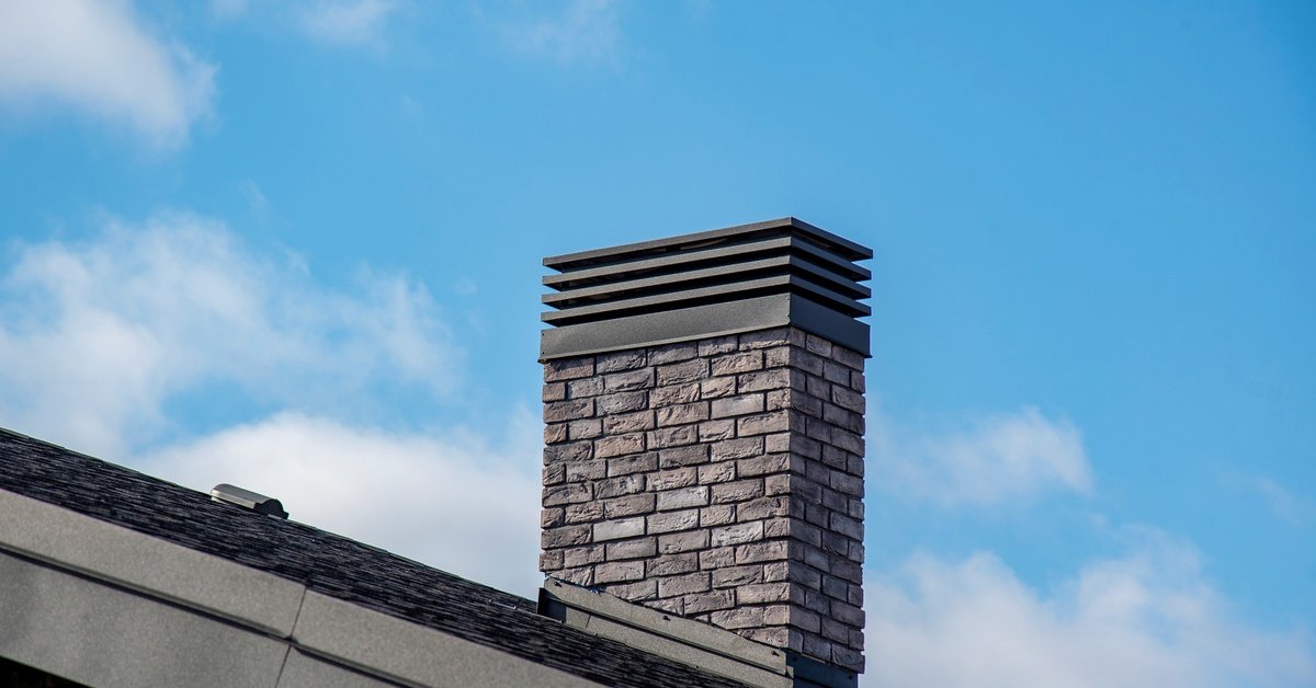 A brick chimney with a black metal cap rising above a sloped roof beneath a bright blue sky with scattered clouds.