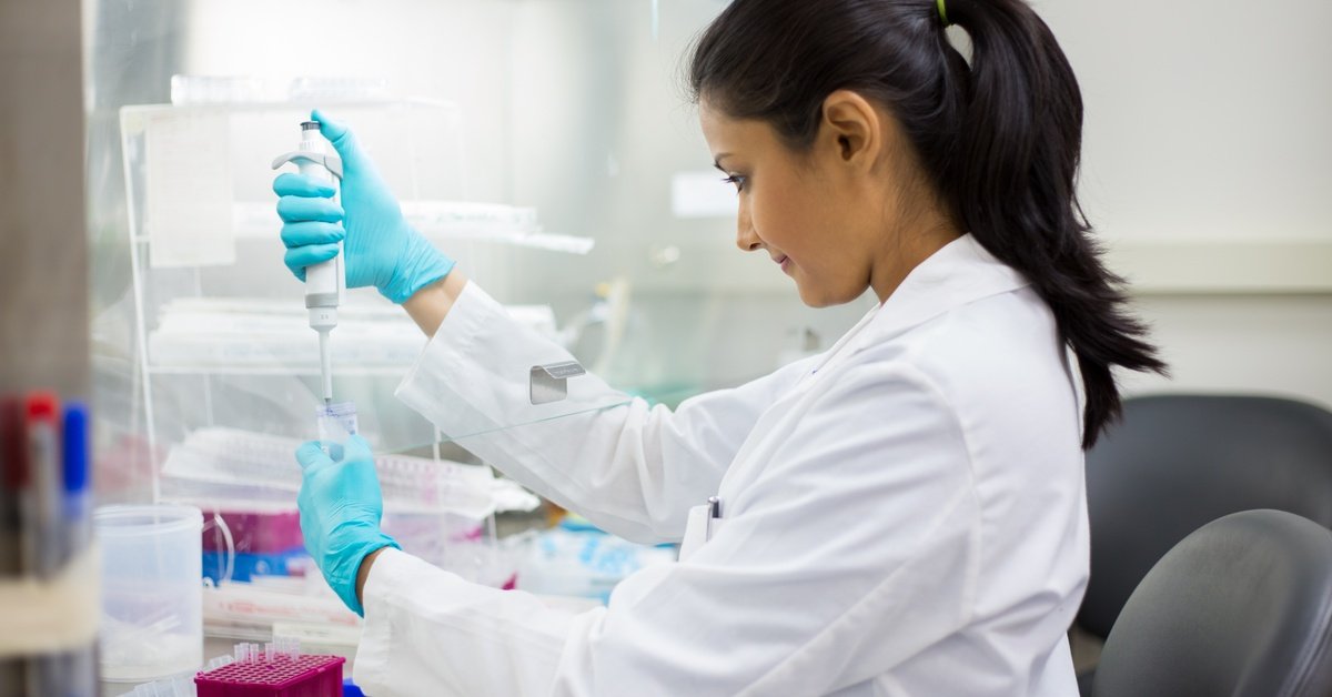 A female scientist sits in a lab wearing a white lab coat and using a scientific tool to drop liquid into a beaker.