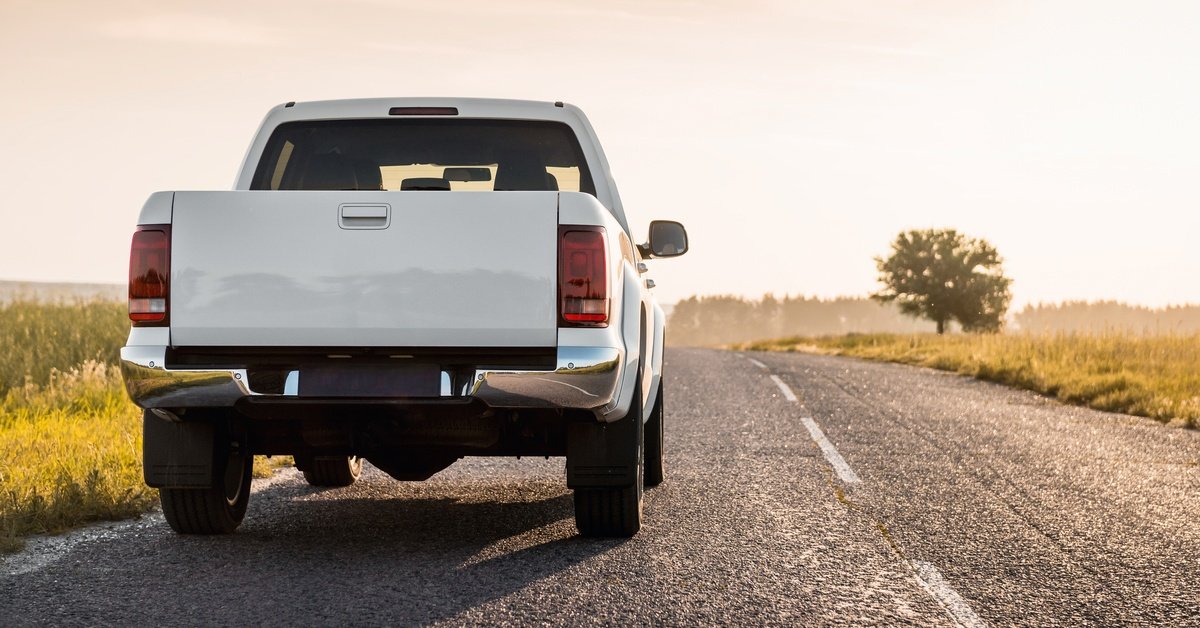 A white pickup truck is driving down the road at dusk. The road is surrounded by farmland, grass, and trees.