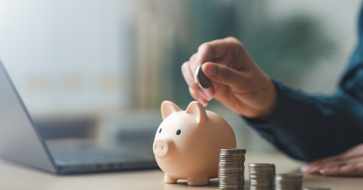A close-up view shows a person holding a coin above a small pink piggy bank. A stack of coins and a laptop is next to the bank.