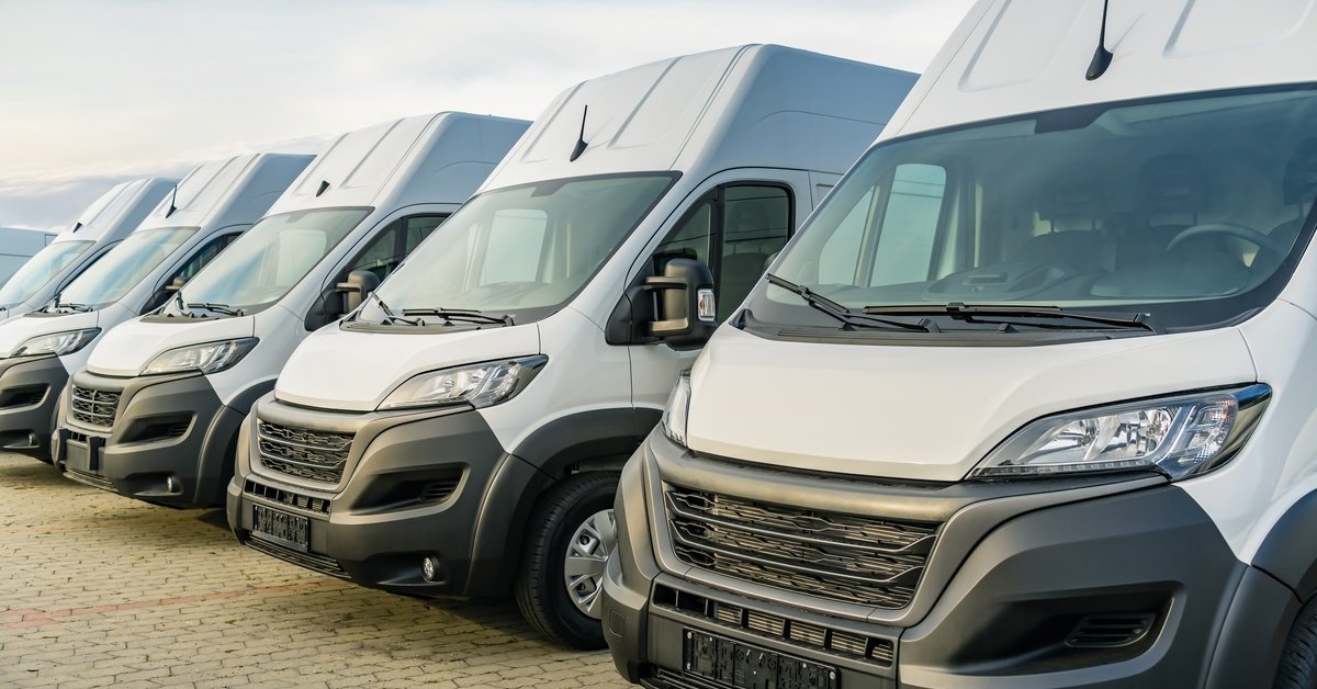 A row of white commercial vans parked side by side on pavement under a partly cloudy sky during daytime.