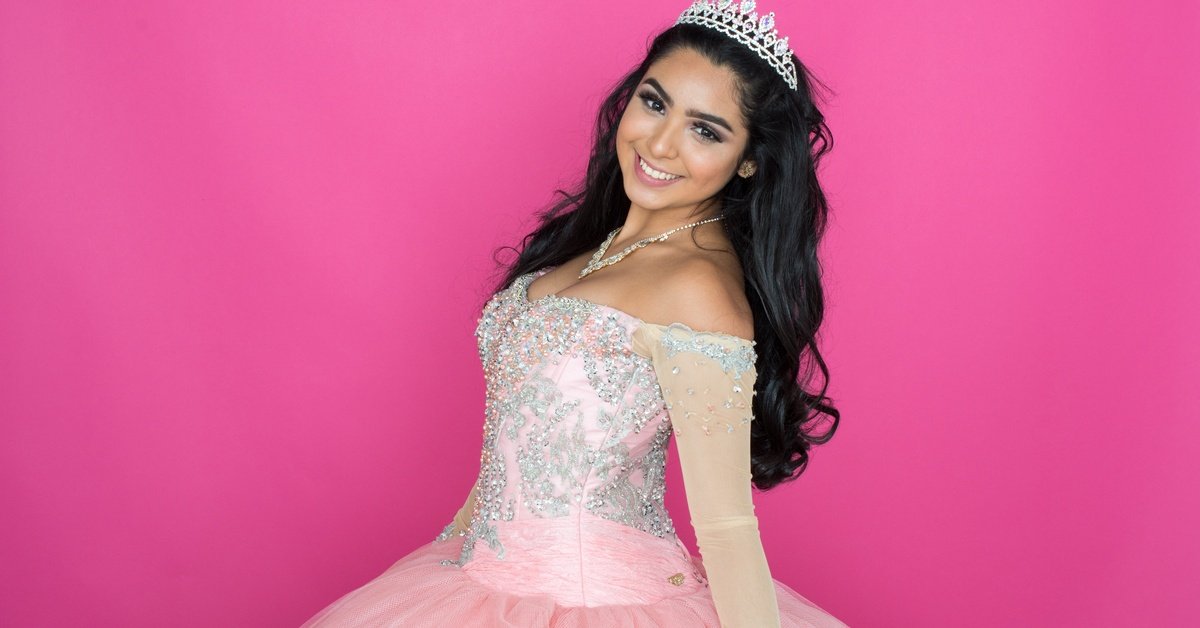 A young woman wearing a silver tiara and a pink embellished dress smiles against a solid pink background.