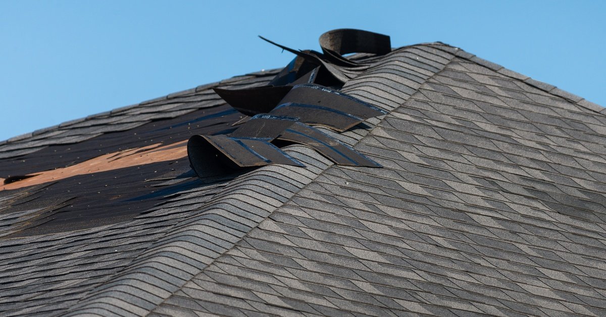 Storm-damaged asphalt shingle roof with torn ridge shingles exposing underlayment beneath a clear blue sky.