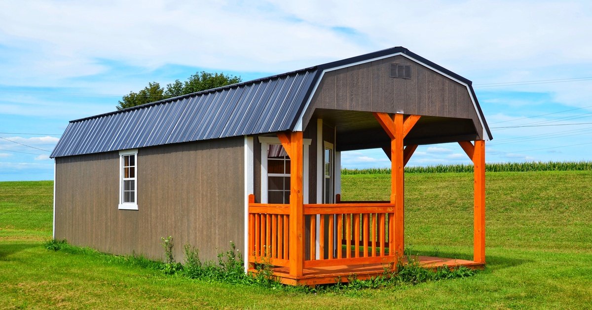 A small barn-style shed with a covered front porch stands on a grassy field beneath a bright blue sky with clouds.