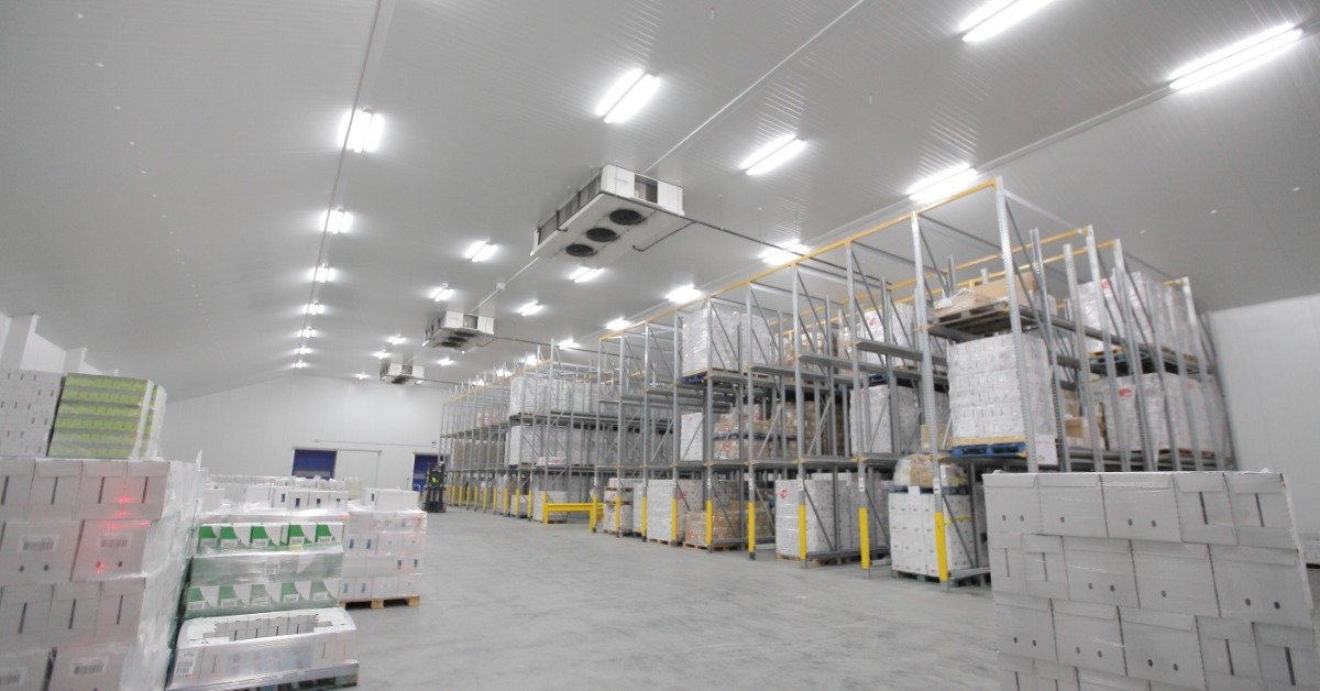A large warehouse stores palletized inventory on tall storage racks beneath bright overhead lighting and cooling units.