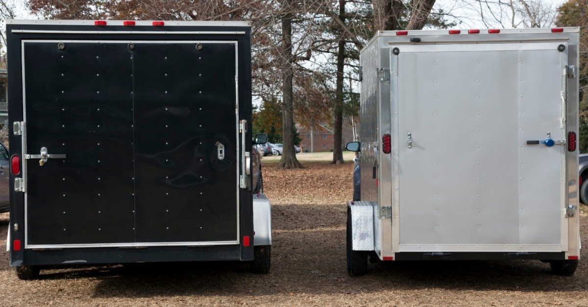 Two enclosed cargo trailers, one black and one silver, parked side by side outdoors with their rear doors closed.
