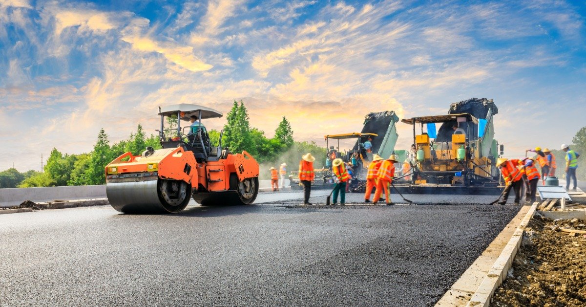 A crew of people wearing orange safety vests is working on repaving a large road. Large machinery is there.