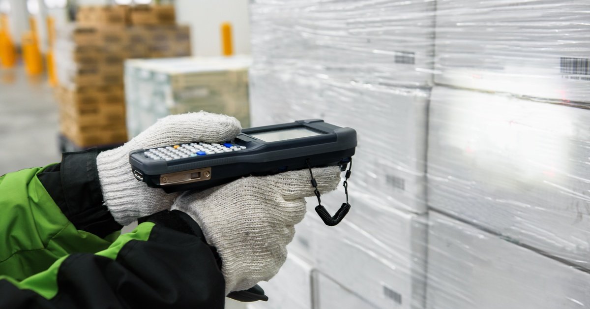A person wearing gloves and a coat is using a handheld barcode scanner to scan a stack of white cardboard boxes.
