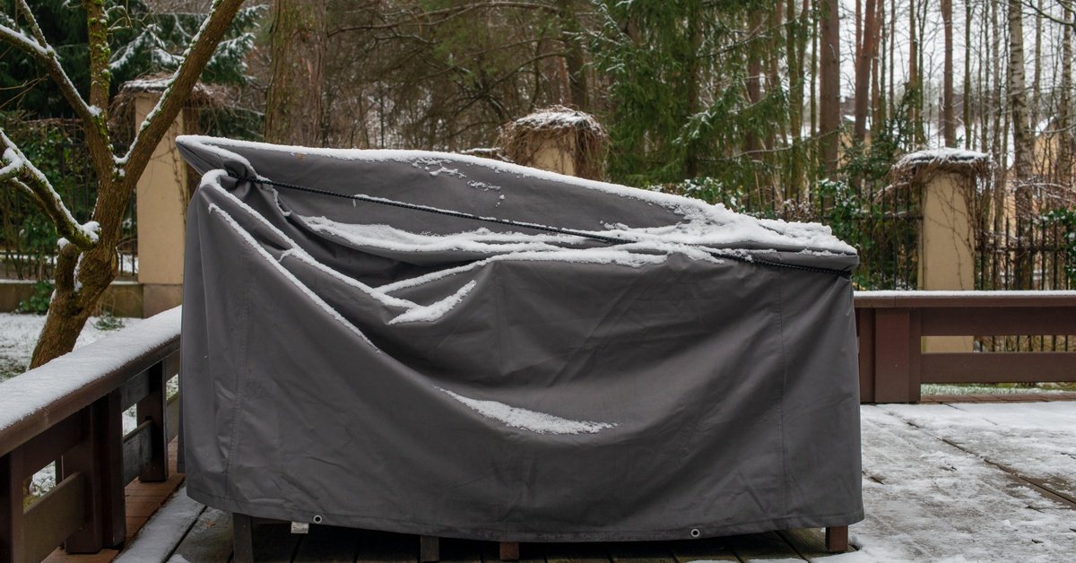 Snow-covered outdoor furniture wrapped in a gray protective cover on a wooden deck surrounded by trees.