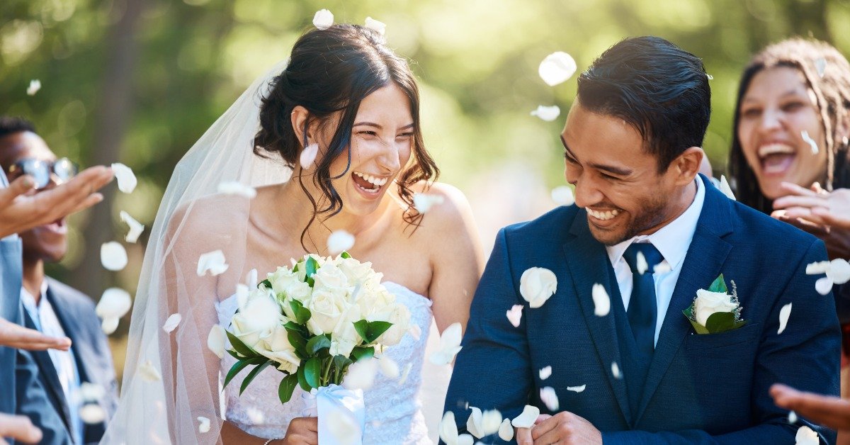 Smiling bride holding white roses and groom in a navy suit celebrate as guests toss white flower petals around them.