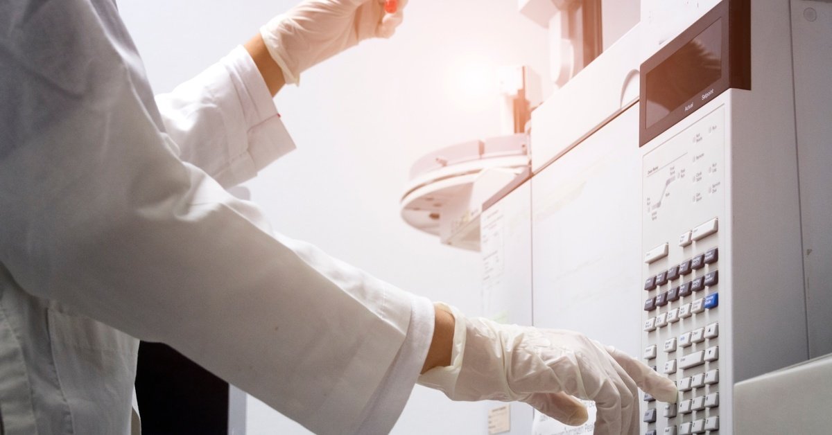 A person wearing a white lab coat and white latex gloves is holding a small vial and pressing the button on a machine.