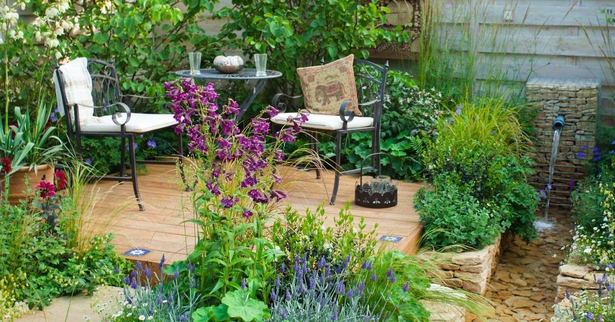 Two outdoor chairs sit next to a circular table on a small wooden deck. The deck is surrounded by flowers.