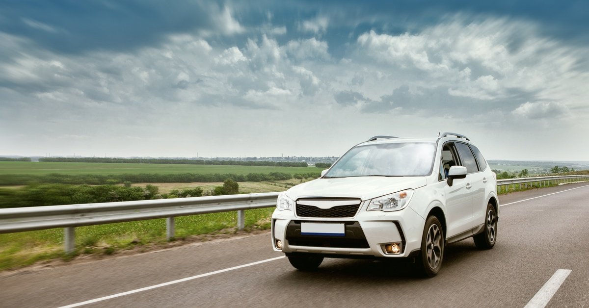 White modern car driving on a long road next to a large field outside. Many large clouds hang overhead.