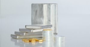 Stacks of silver coins sit in front of a stack of silver bars and a pile of gold bars. The background is white.