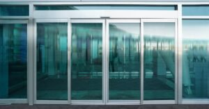 A wide automatic door entryway in the front of a business's lobby, with the interior obscured by the blue-green glass.