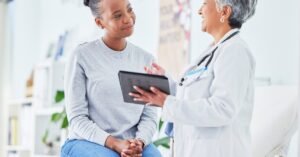 Physician reviews information on tablet with seated patient, both engaged in conversation in clean medical office.