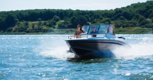 A white and blue motorboat moves across a lake with three people on board. Green wooded hills line the background.