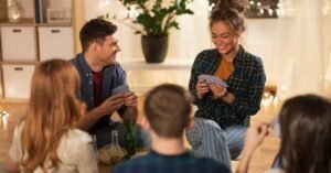 Several people hold playing cards around a low coffee table topped with drinks. Warm string lights light the room.