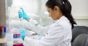 A female scientist sits in a lab wearing a white lab coat and using a scientific tool to drop liquid into a beaker.