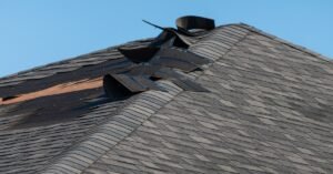 Storm-damaged asphalt shingle roof with torn ridge shingles exposing underlayment beneath a clear blue sky.